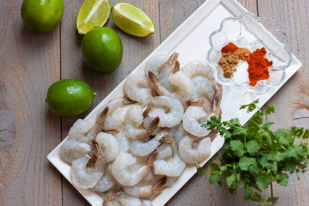 A platter of raw peeled and deveined shrimp next to a bowl of spices, a bunch of french cilantro, and several limes and lime wedges.