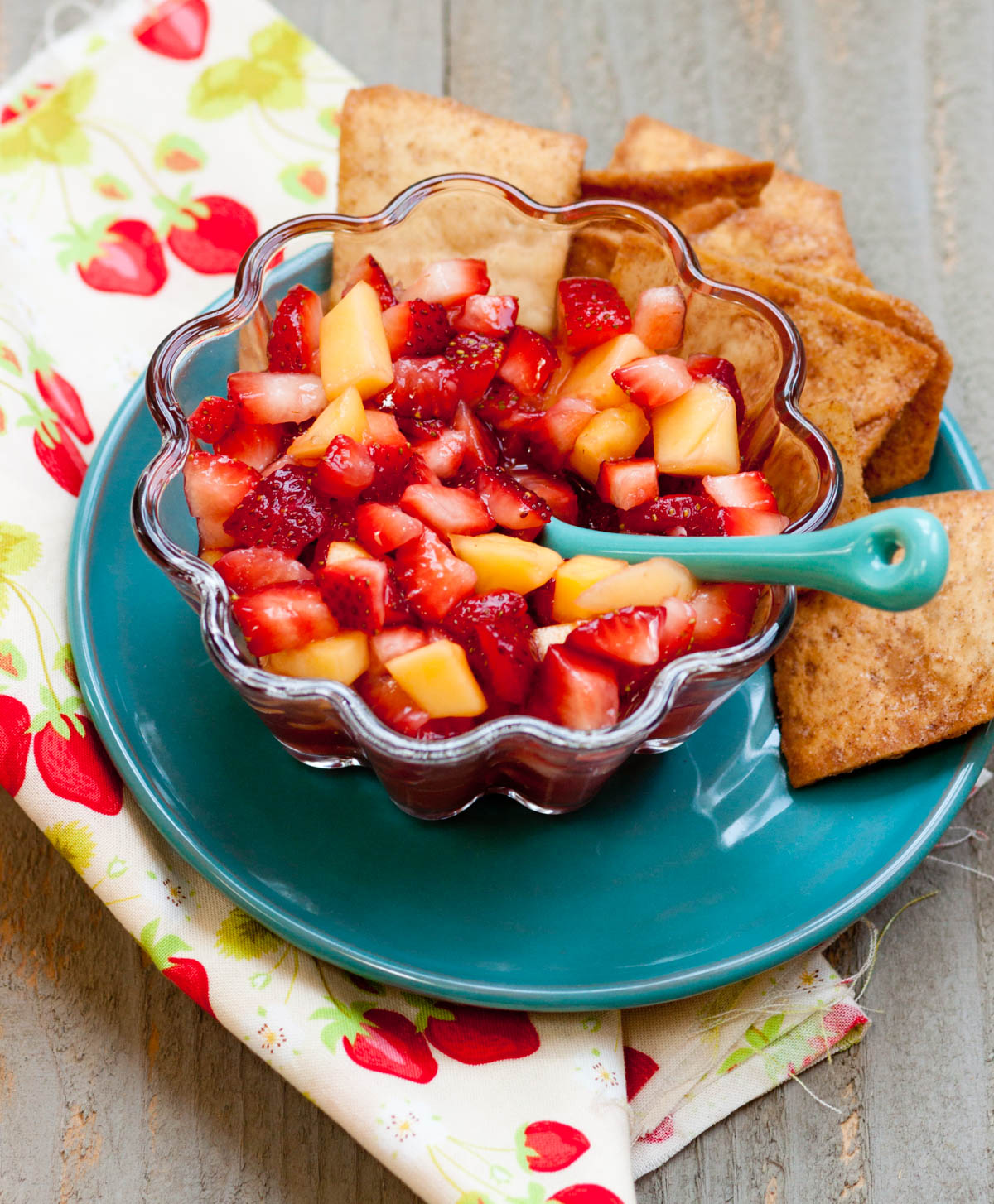 A bowl of strawberry mango salsa has a blue spoon and cinnamon pita chips on the plate next to it.