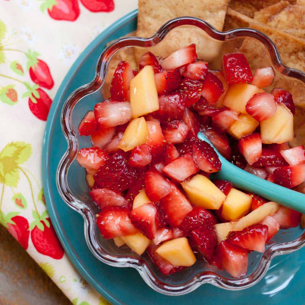 A bowl of strawberry mango salsa has a blue spoon and cinnamon pita chips on the plate next to it.