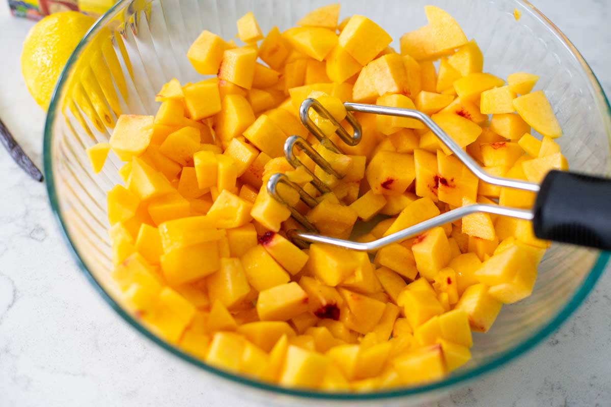Peeled and chopped fresh peaches are about to be mashed by a potato masher.