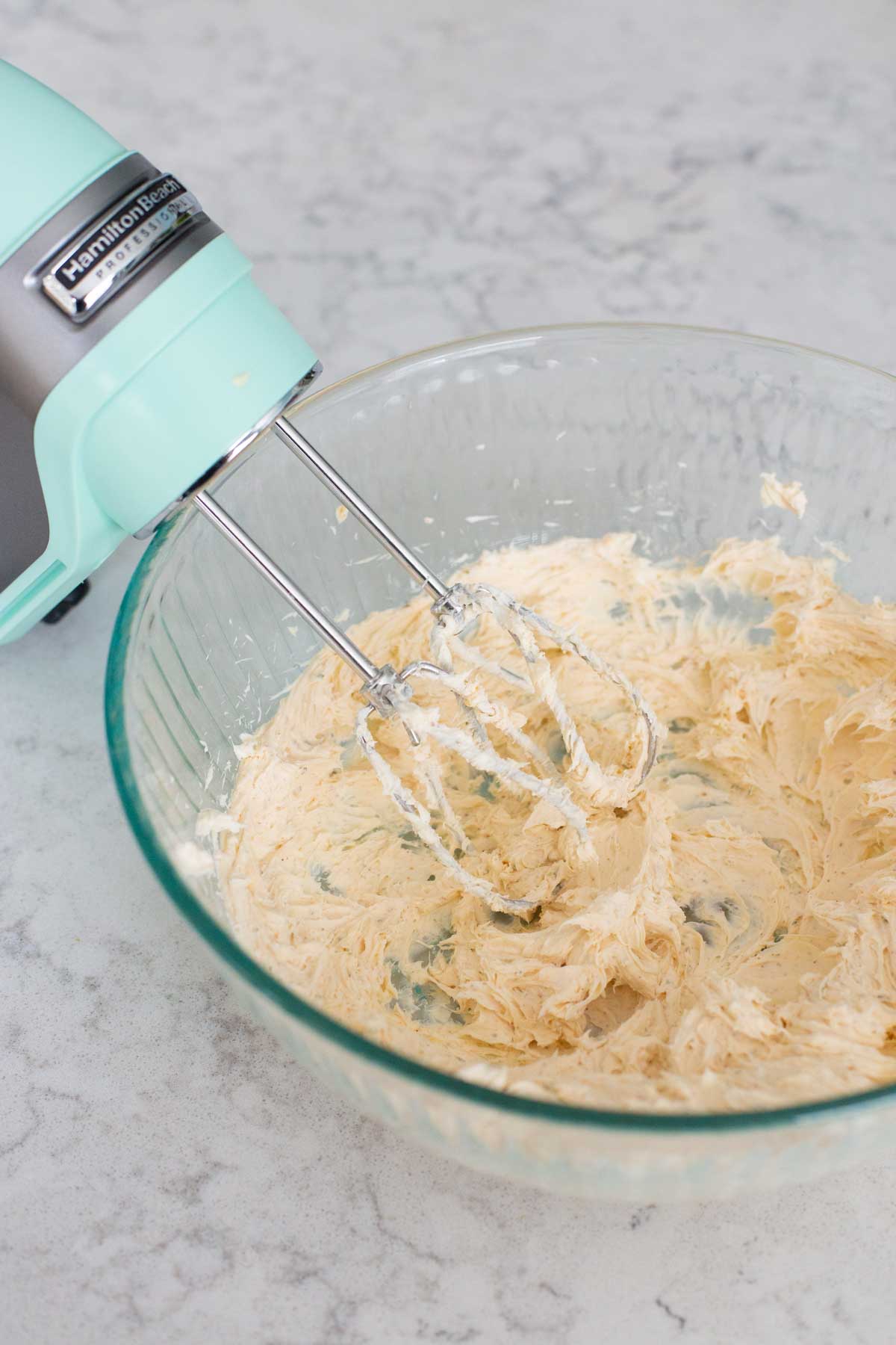 A mixing bowl filled with cream cheese and seasonings is being beaten by a hand mixer.