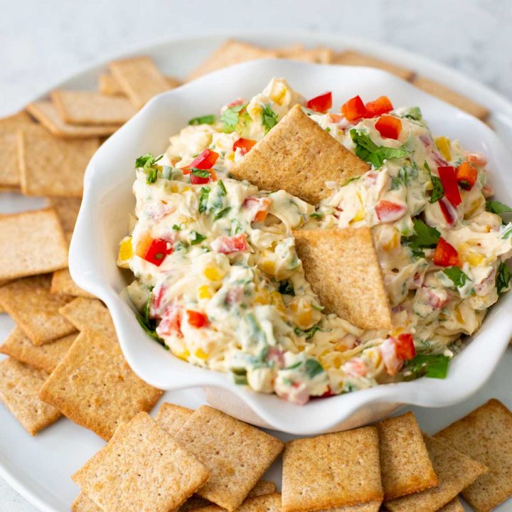 A creamy dip with corn, peppers, and cilantro sits in a bowl surrounded by wheat crackers.