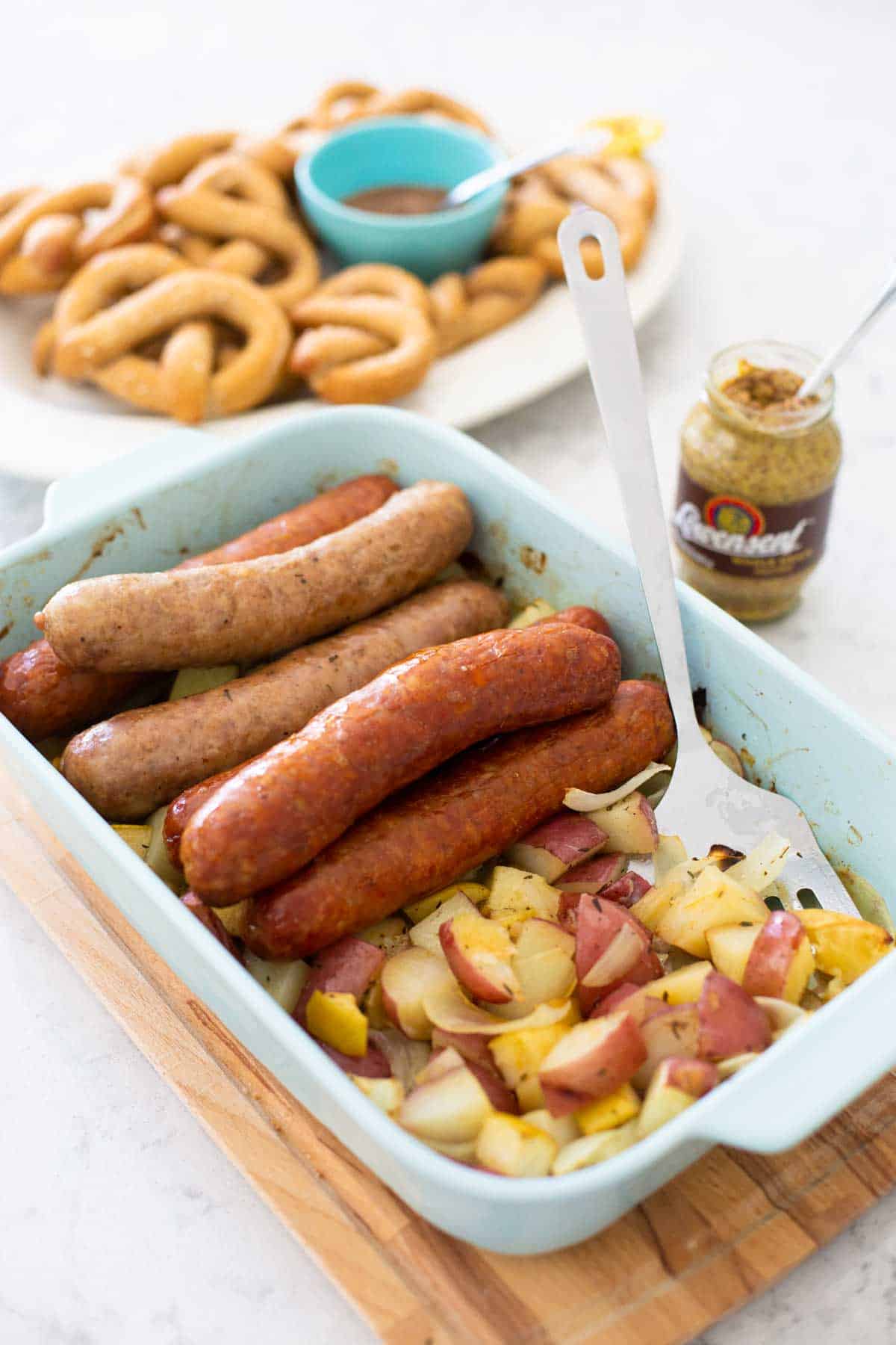 A baking dish is filled with roasted sausages, potatoes, and apples. A jar of mustard and a platter of baked pretzels are in the background.