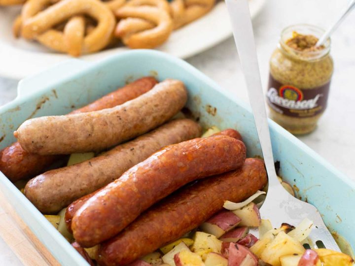 A baking dish is filled with roasted sausages, potatoes, and apples. A jar of mustard and a platter of baked pretzels are in the background.