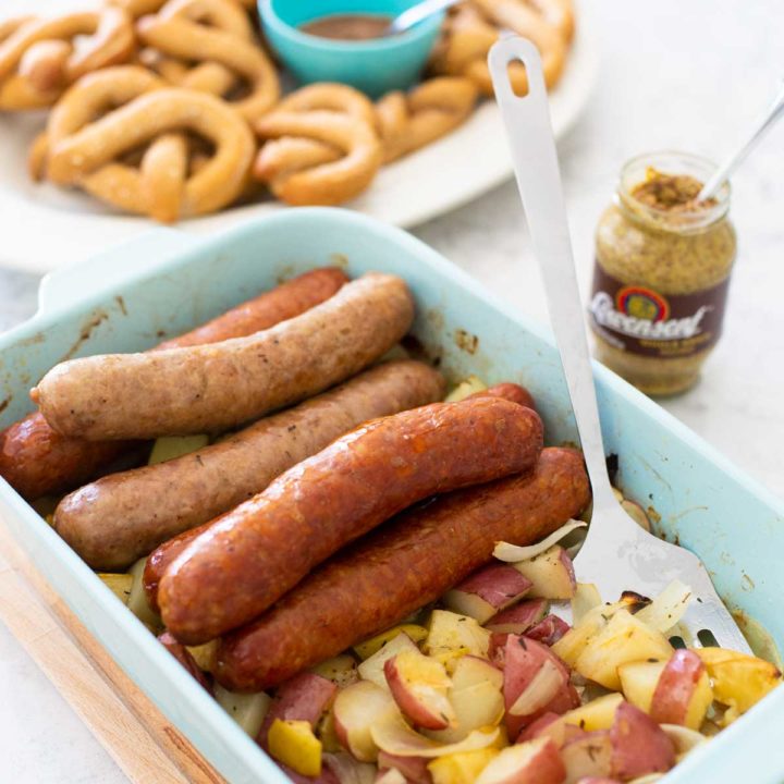 A baking dish is filled with roasted sausages, potatoes, and apples. A jar of mustard and a platter of baked pretzels are in the background.