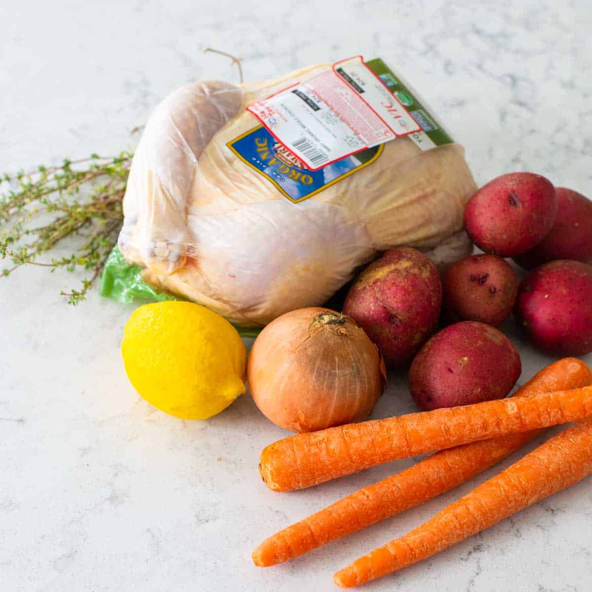 The chicken, potatoes, carrots, onion, lemon, and thyme are sitting on the kitchen counter.