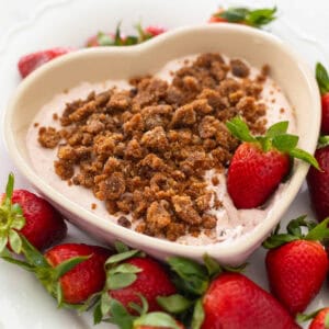 A heart shaped dish of strawberry cheesecake dip has cookie crumbles on top and is surrounded by fresh strawberries for dunking.