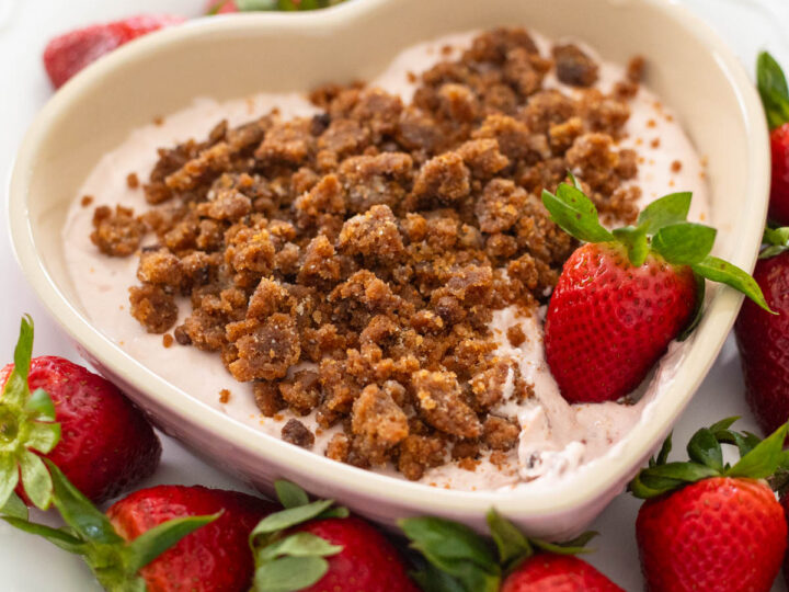 A heart shaped dish of strawberry cheesecake dip has cookie crumbles on top and is surrounded by fresh strawberries for dunking.
