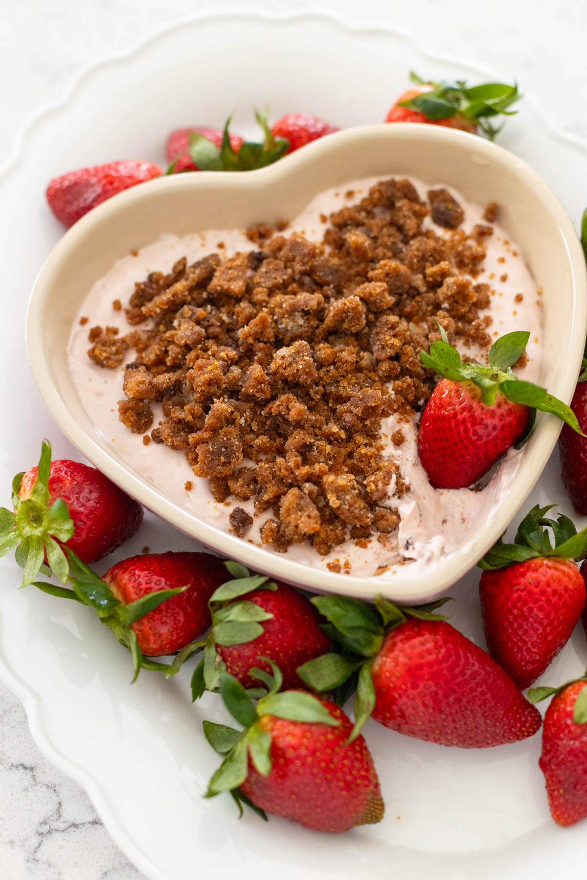 A heart shaped dish of strawberry cheesecake dip has cookie crumbles on top and is surrounded by fresh strawberries for dunking.