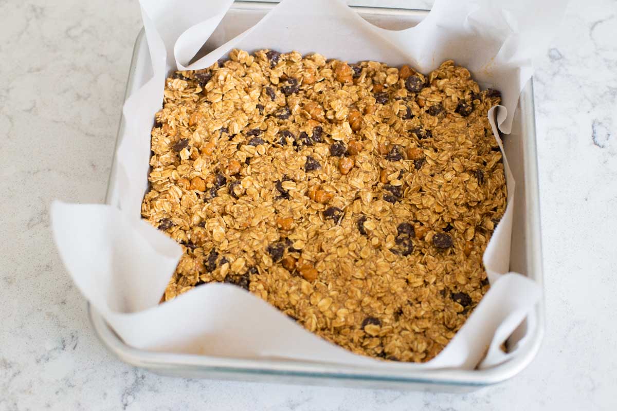 An 8x8-inch pan has parchment paper and the granola bar dough patted into place.