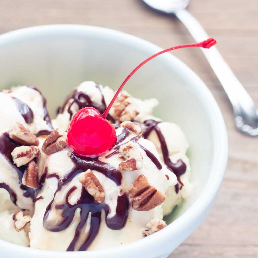 A bowl filled with a homemade turtle sundae: vanilla ice cream drizzled with hot fudge, caramel, sprinkled with salted pecans, and a red cherry on top.