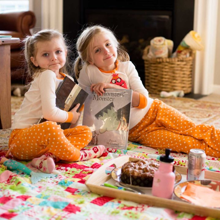 Two girls in Halloween jammies hold Halloween picture books and are sitting on the living room floor with breakfast treats.