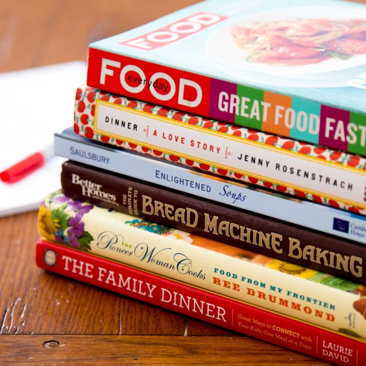 A stack of colorful cookbooks sits next to a pad of paper with a pen.