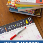 A meal planning pad with a pen sits next to a stack of colorful cookbooks.