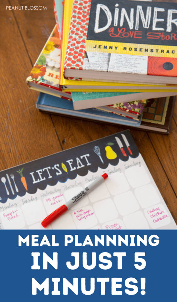 A meal planning pad with a pen sits next to a stack of colorful cookbooks.