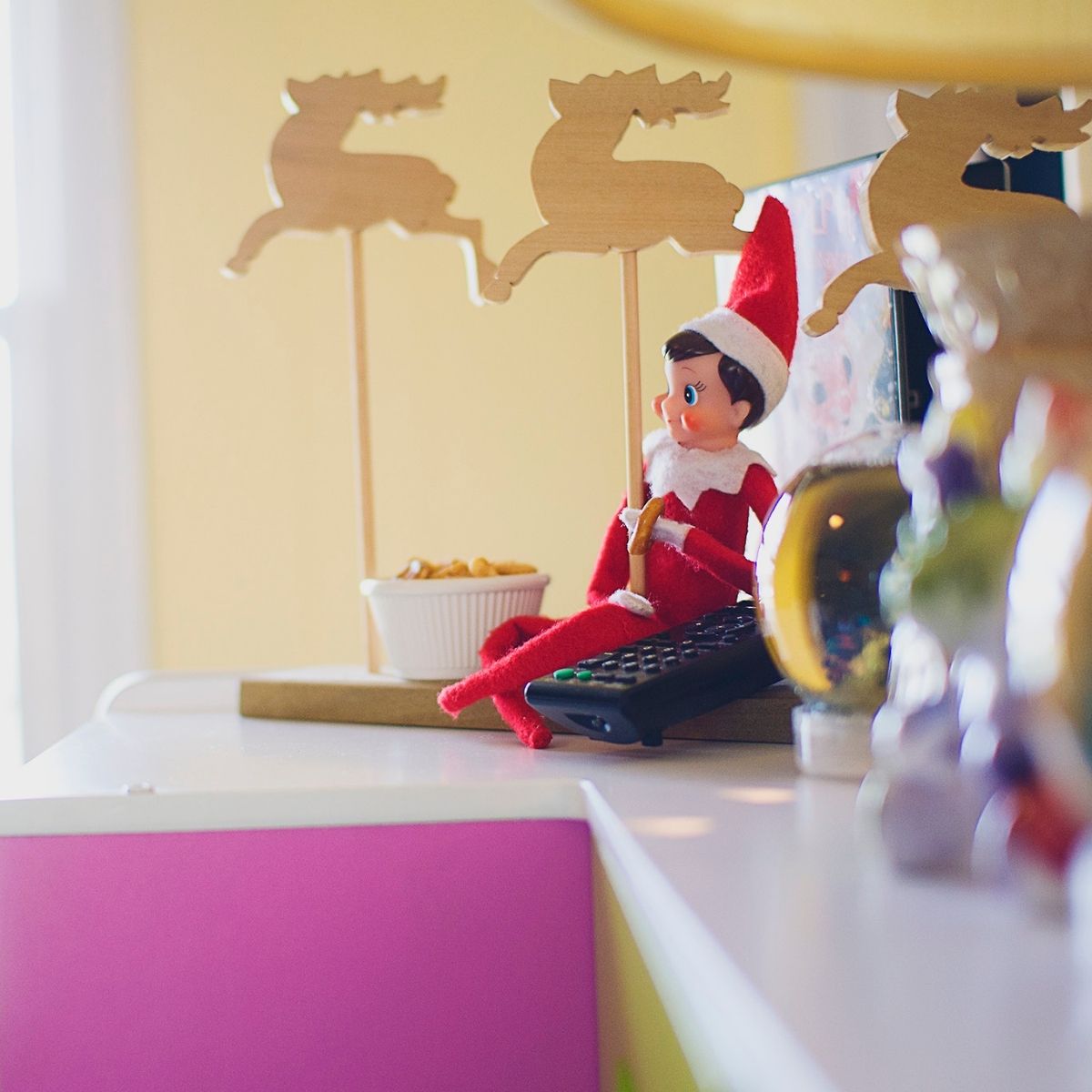An Elf on the Shelf sits by the tv remote on a shelf.