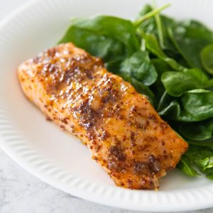 The salmon filet is coated in a brown sugar and mustard glaze and served on a white plate next to a bed of baby spinach.