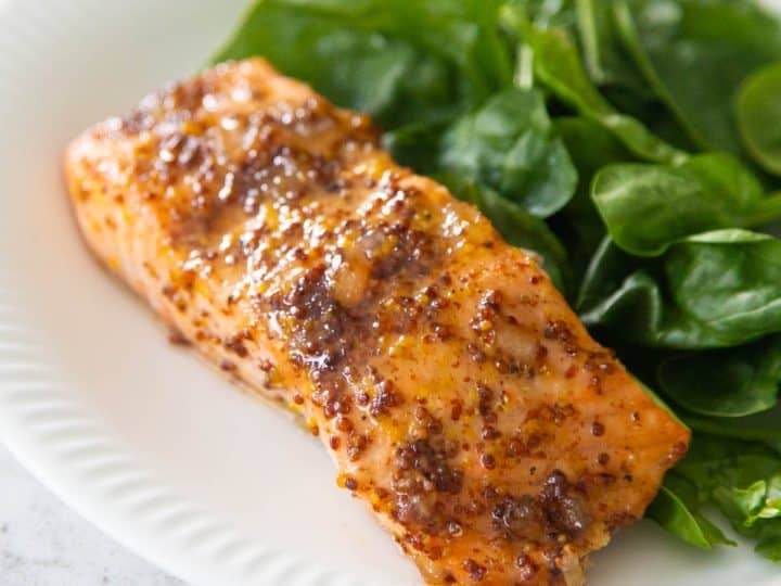 The salmon filet is coated in a brown sugar and mustard glaze and served on a white plate next to a bed of baby spinach.