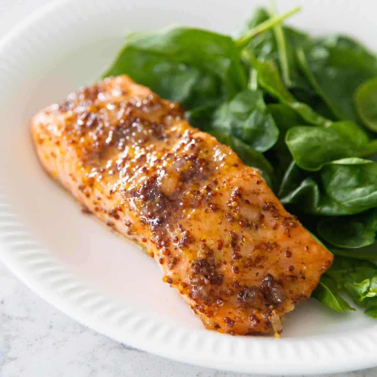 The salmon filet is coated in a brown sugar and mustard glaze and served on a white plate next to a bed of baby spinach.