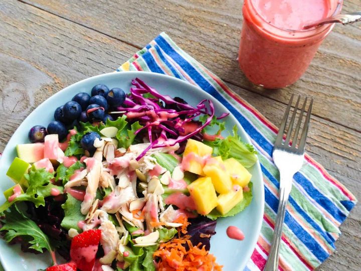A jar of homemade strawberry vinaigrette sits next to a plate full of rainbow salad with fresh strawberries, carrots, pineapple, purple cabbage, blueberries, chicken, and lettuce.