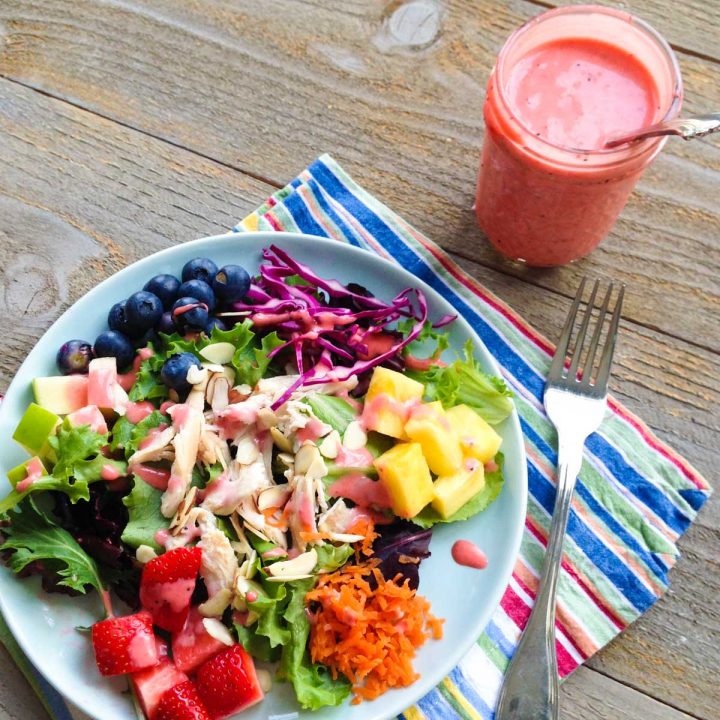A jar of homemade strawberry vinaigrette sits next to a plate full of rainbow salad with fresh strawberries, carrots, pineapple, purple cabbage, blueberries, chicken, and lettuce.