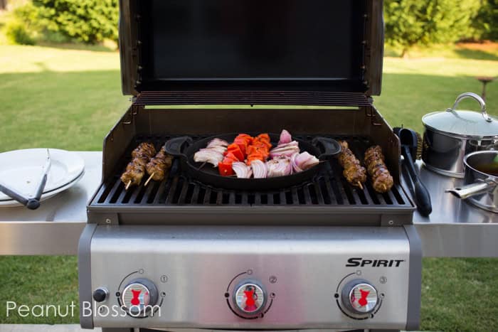 A grill with the lid open has a tray of vegetables and 4 skewers of meatball kabobs.