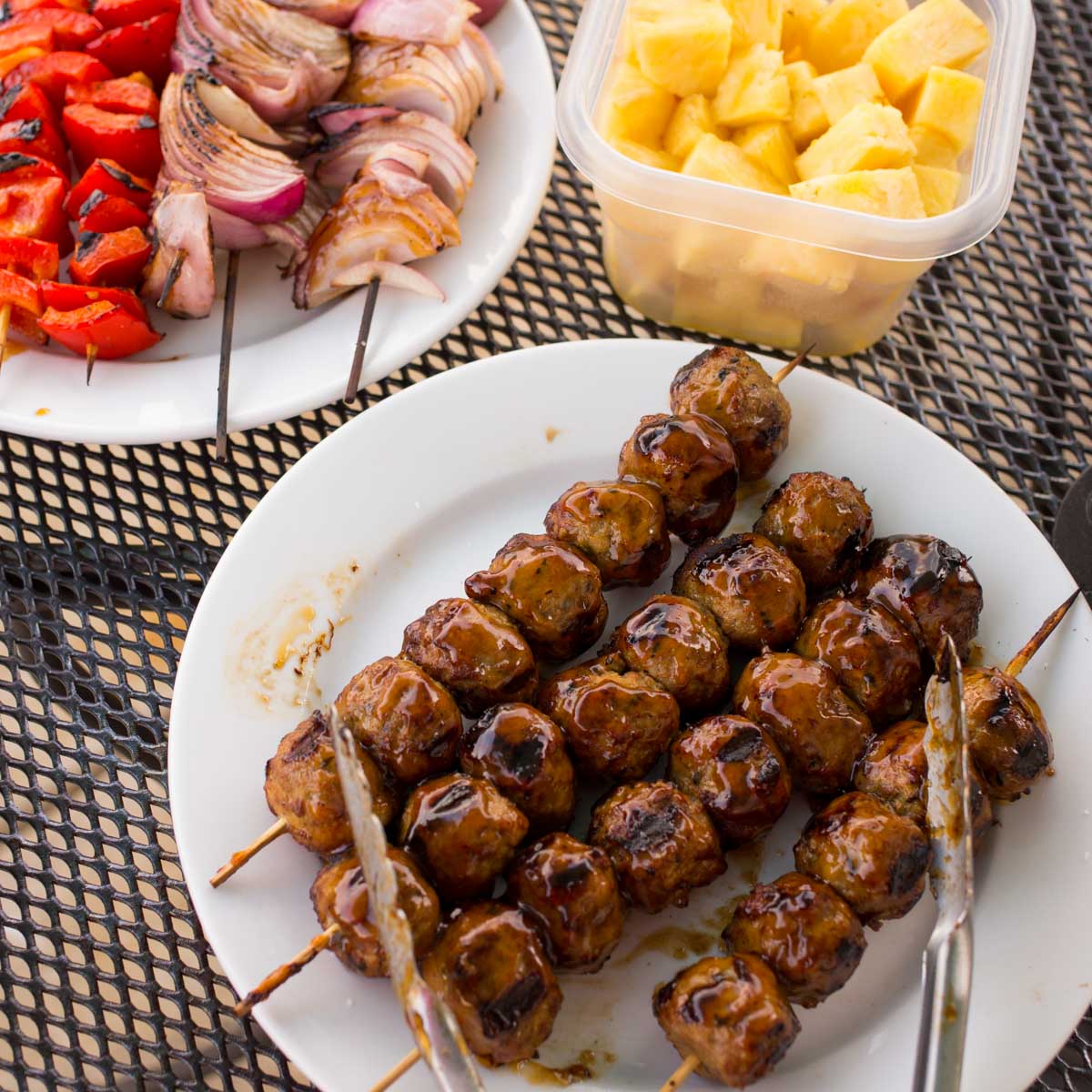 A platter of 4 meatball skewers with teriyaki glaze. A plate of grilled vegetables and fresh pineapple are in the back.