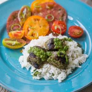 A blue plate holds a serving of rice with chunks of steak drizzled with fresh green chimichurri sauce. Fresh tomatoes are sliced in the back.