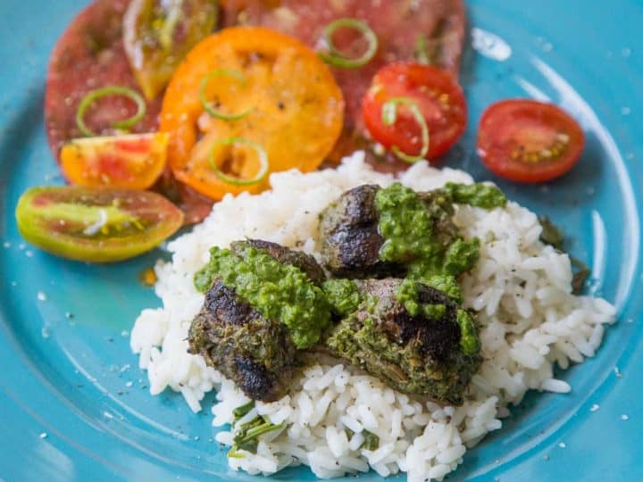 A blue plate holds a serving of rice with chunks of steak drizzled with fresh green chimichurri sauce. Fresh tomatoes are sliced in the back.