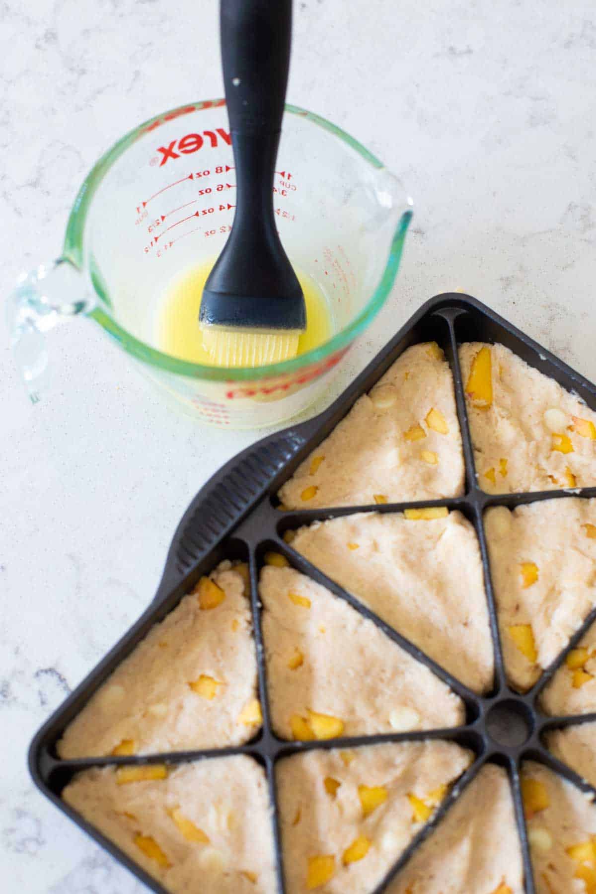 A cup of melted butter with a pastry brush is ready to brush butter on the scones in the baking pan.