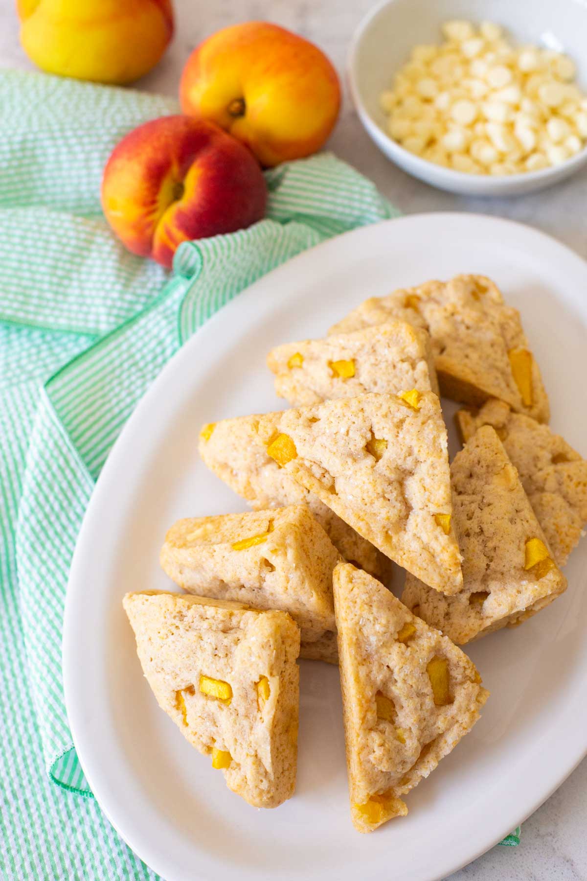 A platter of peach scones has fresh peaches and a bowl of white chocolate chips in the backdrop.