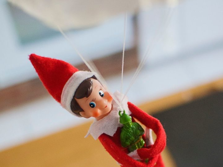 An Elf on the Shelf parachutes with a green army man toy in his arms.