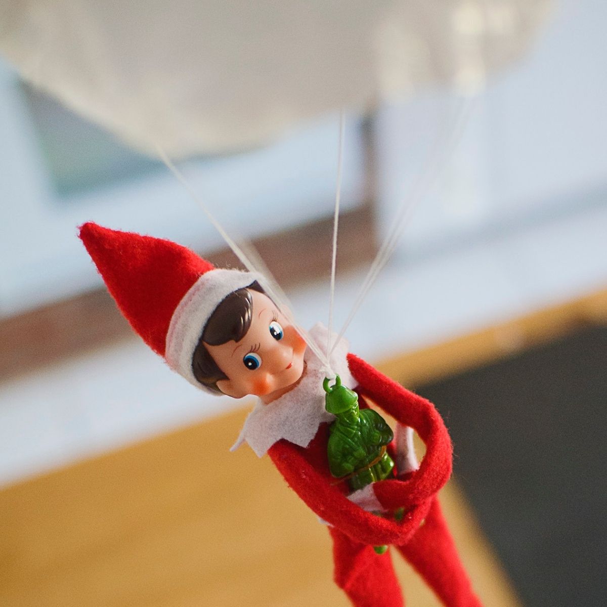 An Elf on the Shelf parachutes with a green army man toy in his arms.