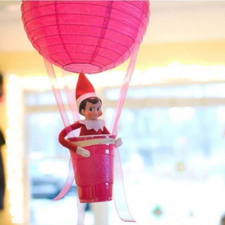 The Elf on the Shelf is sitting in a homemade 'hot air balloon' made from a red plastic cup and a streamer.