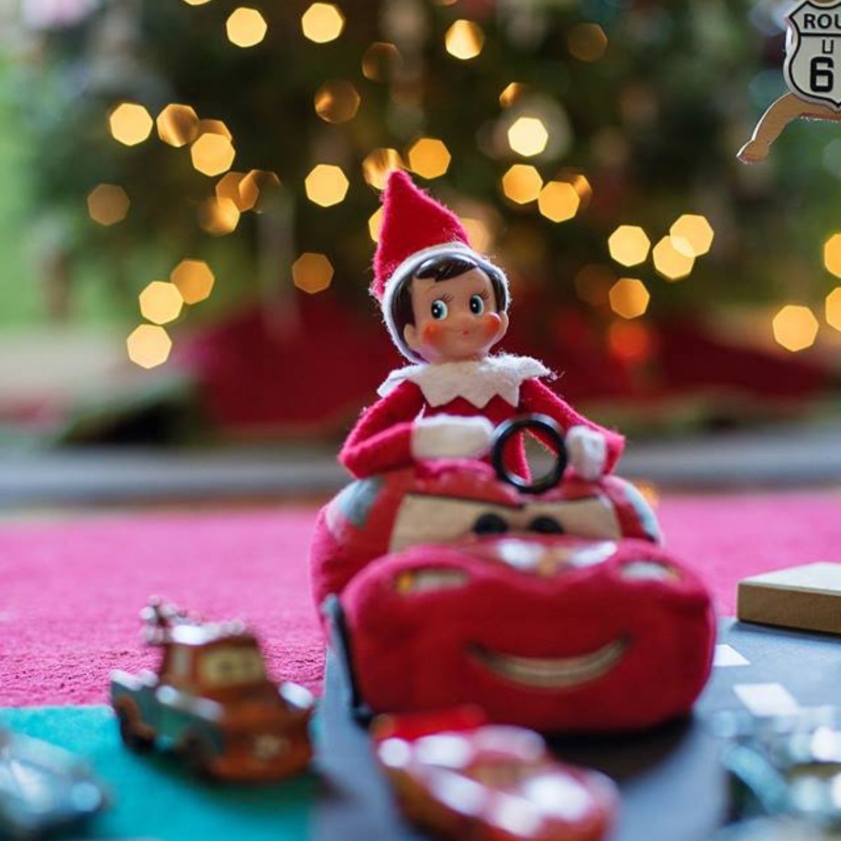 Elf on the Shelf riding a Lightning McQueen car toy with a Christmas tree in the background.