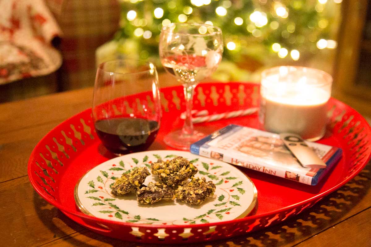 A red tray with treats and two glasses of wine and a lit candle sits on a coffee table in front of a Christmas tree.