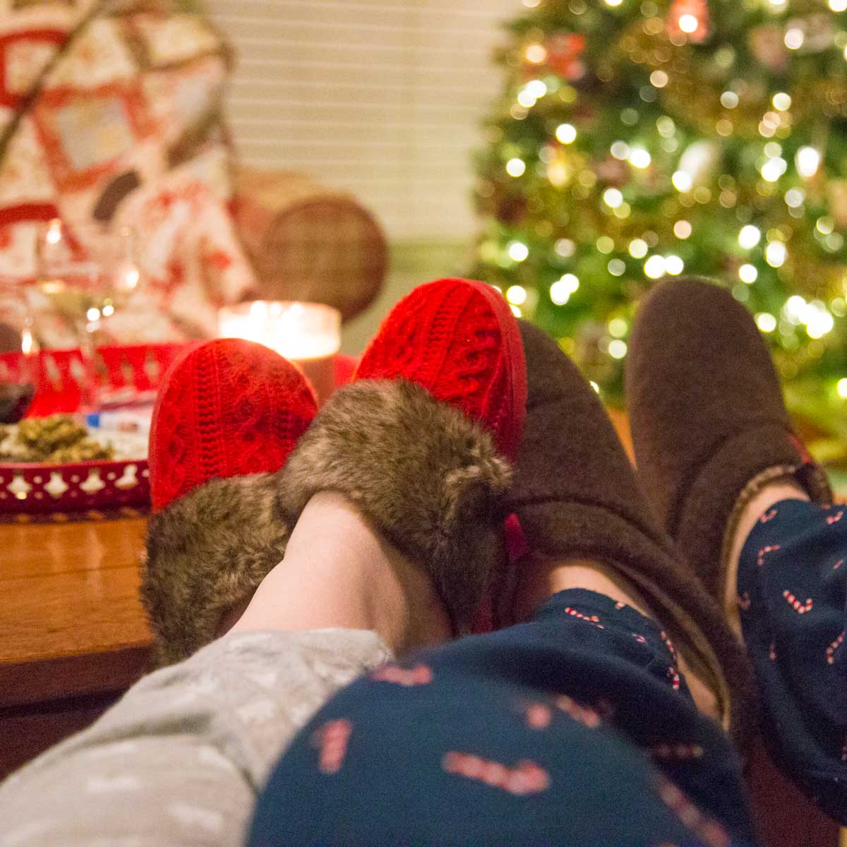 A husband and wife wear Christmas slippers and sit in front of a Christmas tree.