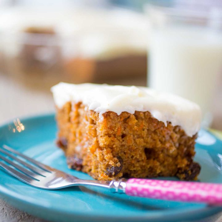 A square slice of carrot cake with cream cheese frosting sits on a blue plate with a pink fork.