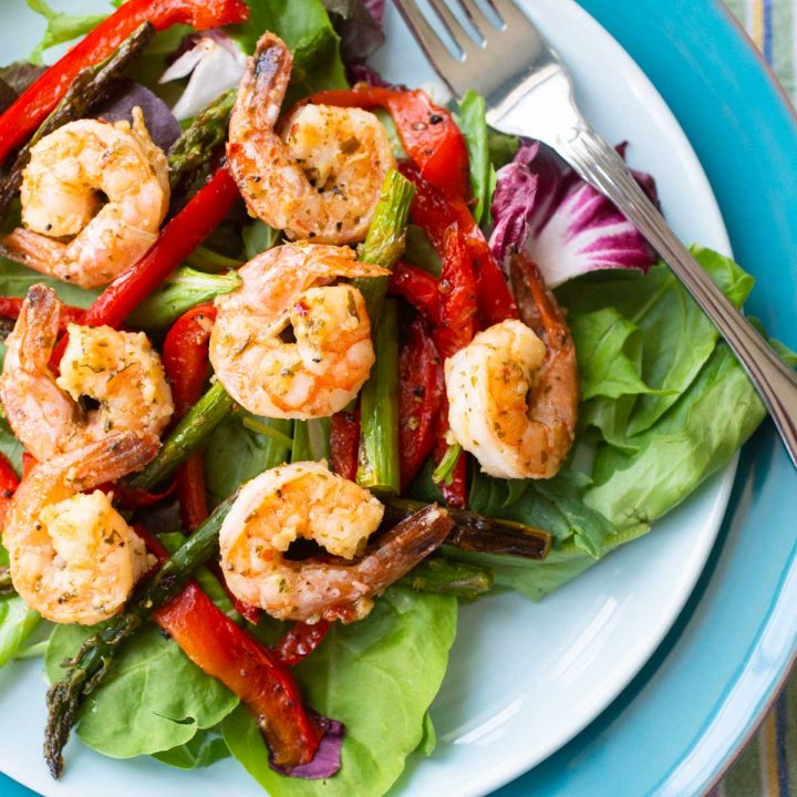 A blue plate has a bed of lettuce topped with lemon garlic shrimp and roasted red peppers and asparagus spears.