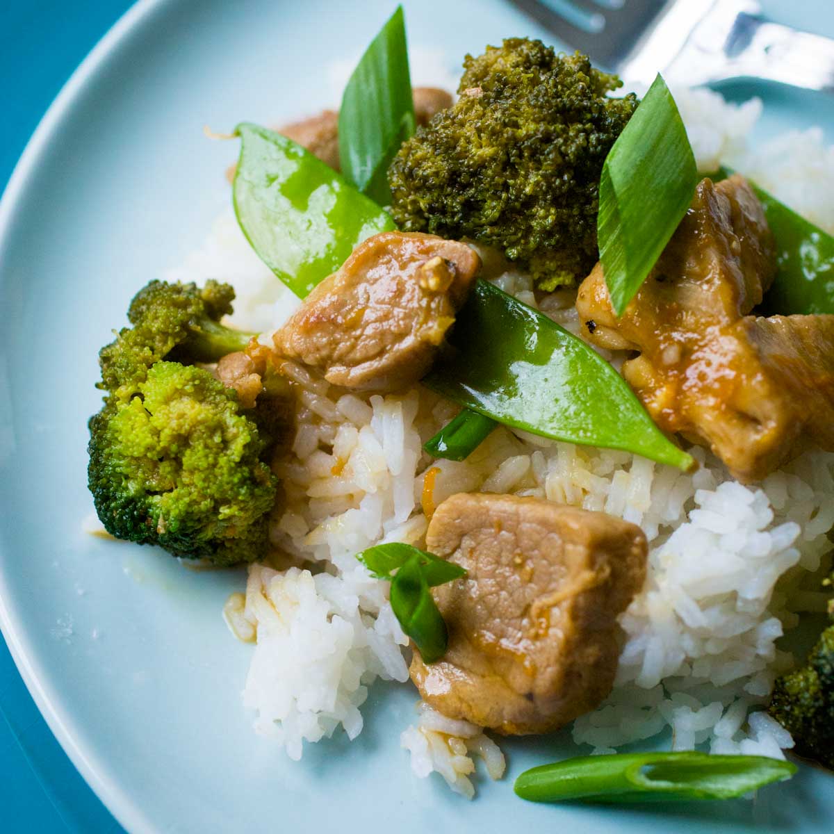 A blue plate has a bed of white rice and is topped with chunks of pork, broccoli spears, pea pods, and a sprinkle of green onions.