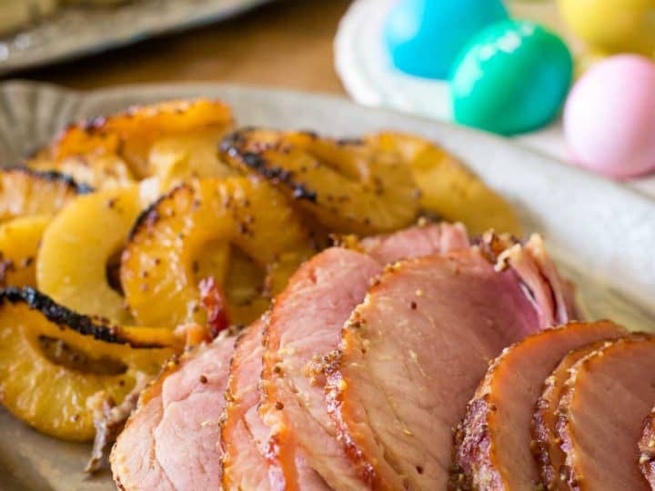 Slices of pineapple glazed ham sits on a platter next to the roasted pineapple slices. A tray of colorful Easter eggs sits in the background.
