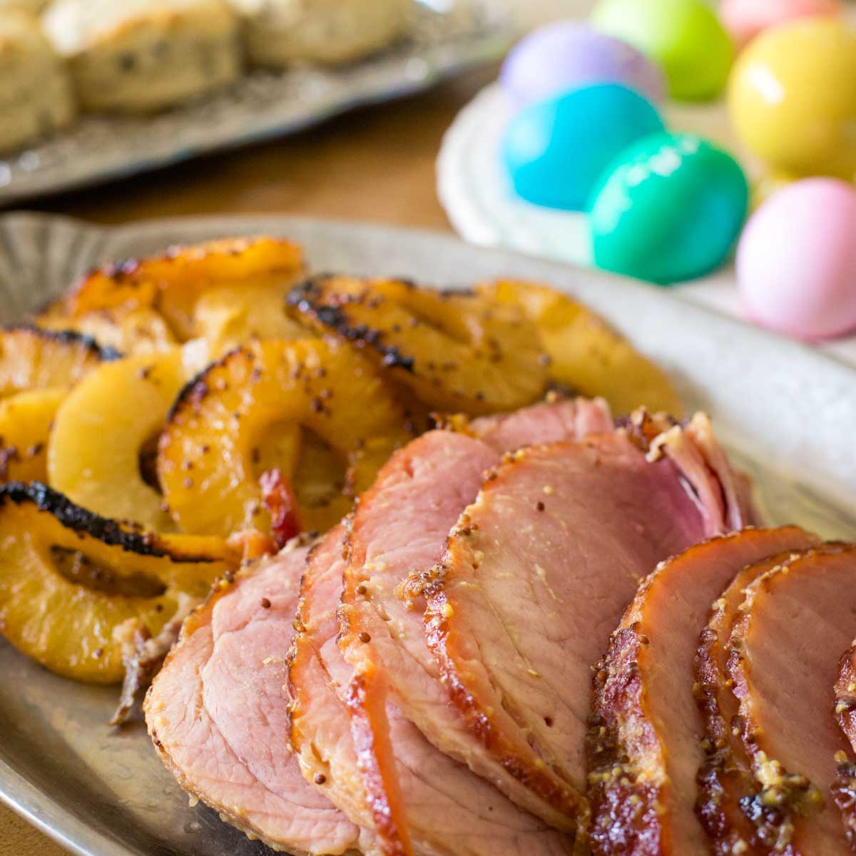 Slices of pineapple glazed ham sits on a platter next to the roasted pineapple slices. A tray of colorful Easter eggs sits in the background.