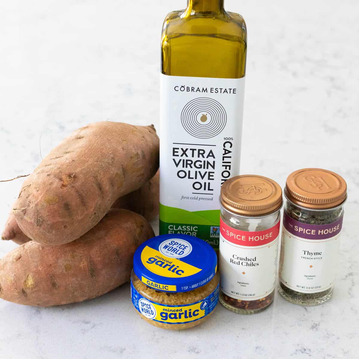 Two fresh sweet potatoes are on the counter next to a bottle of olive oil and some seasonings.