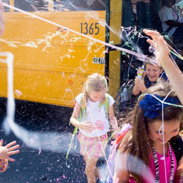 Kids coming off a school bus are getting sprayed with silly string by their parents.