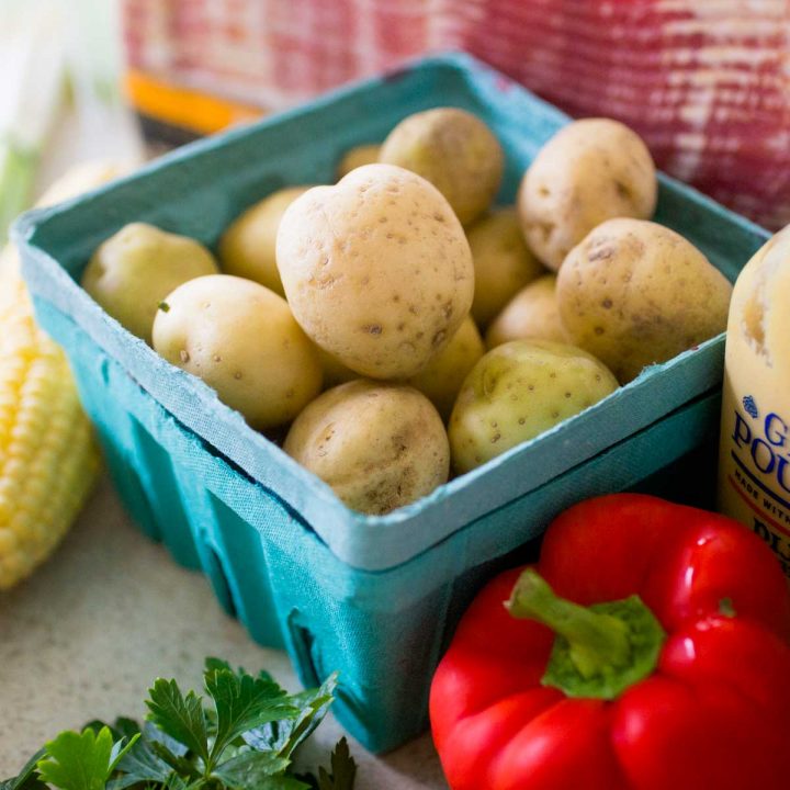 A blue farmer's market carton holds little white potatoes next to a red pepper, ear of corn, and package of bacon.