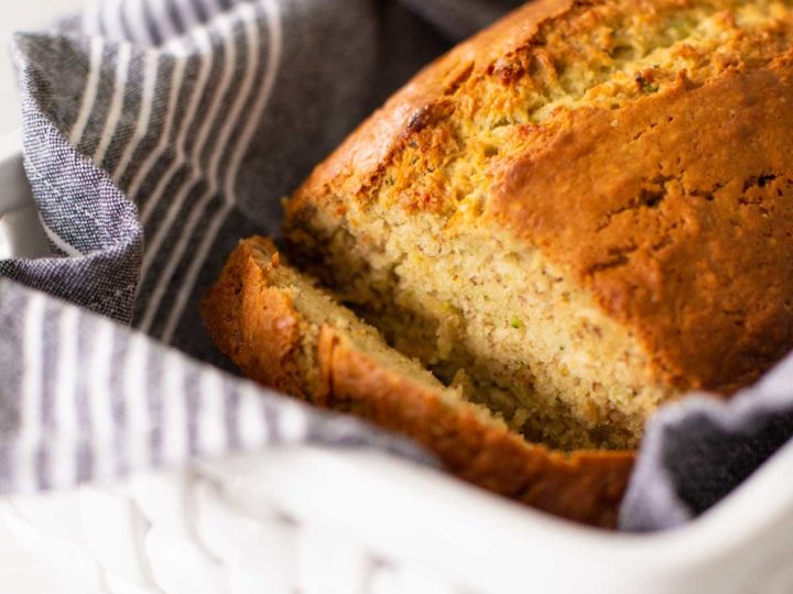 Homemade banana zucchini bread has been sliced to show the inside and sits in a white ceramic bread basket with a grey striped napkin.