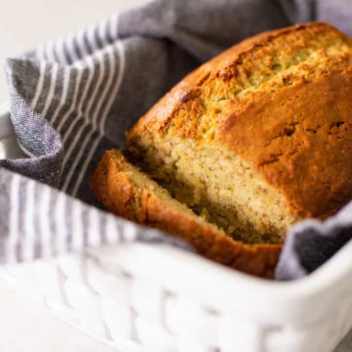 Homemade banana zucchini bread has been sliced to show the inside and sits in a white ceramic bread basket with a grey striped napkin.