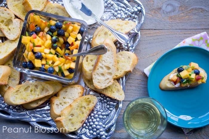 The serving platter has a bowl of peach salsa with blueberries, surrounded by crostini bread. A glass of wine and an appetizer plate with a completed peach bite is off to the side.