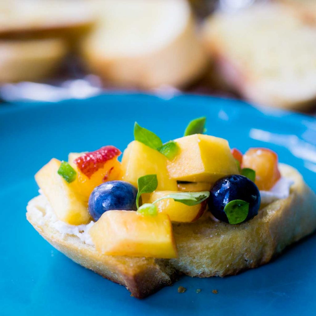 A crostini toast has diced peaches, blueberries, and fresh thyme leaves on a blue plate.