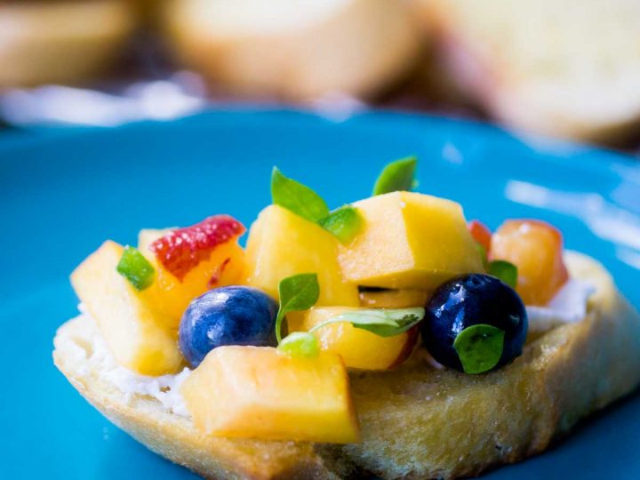 A crostini toast has diced peaches, blueberries, and fresh thyme leaves on a blue plate.